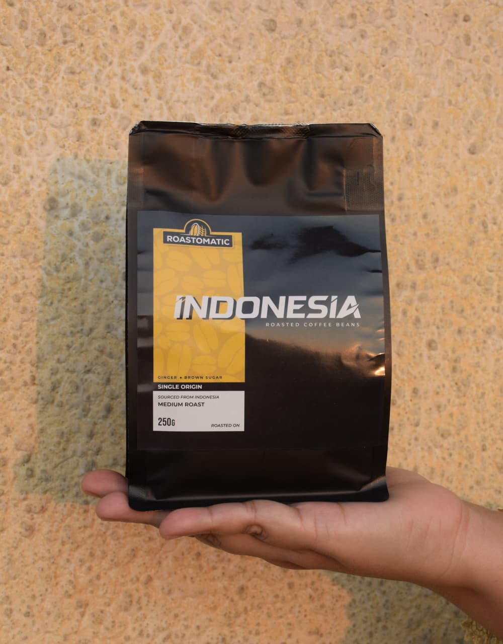 Hand holding a Roastomatic Indonesia coffee bag against a textured beige background