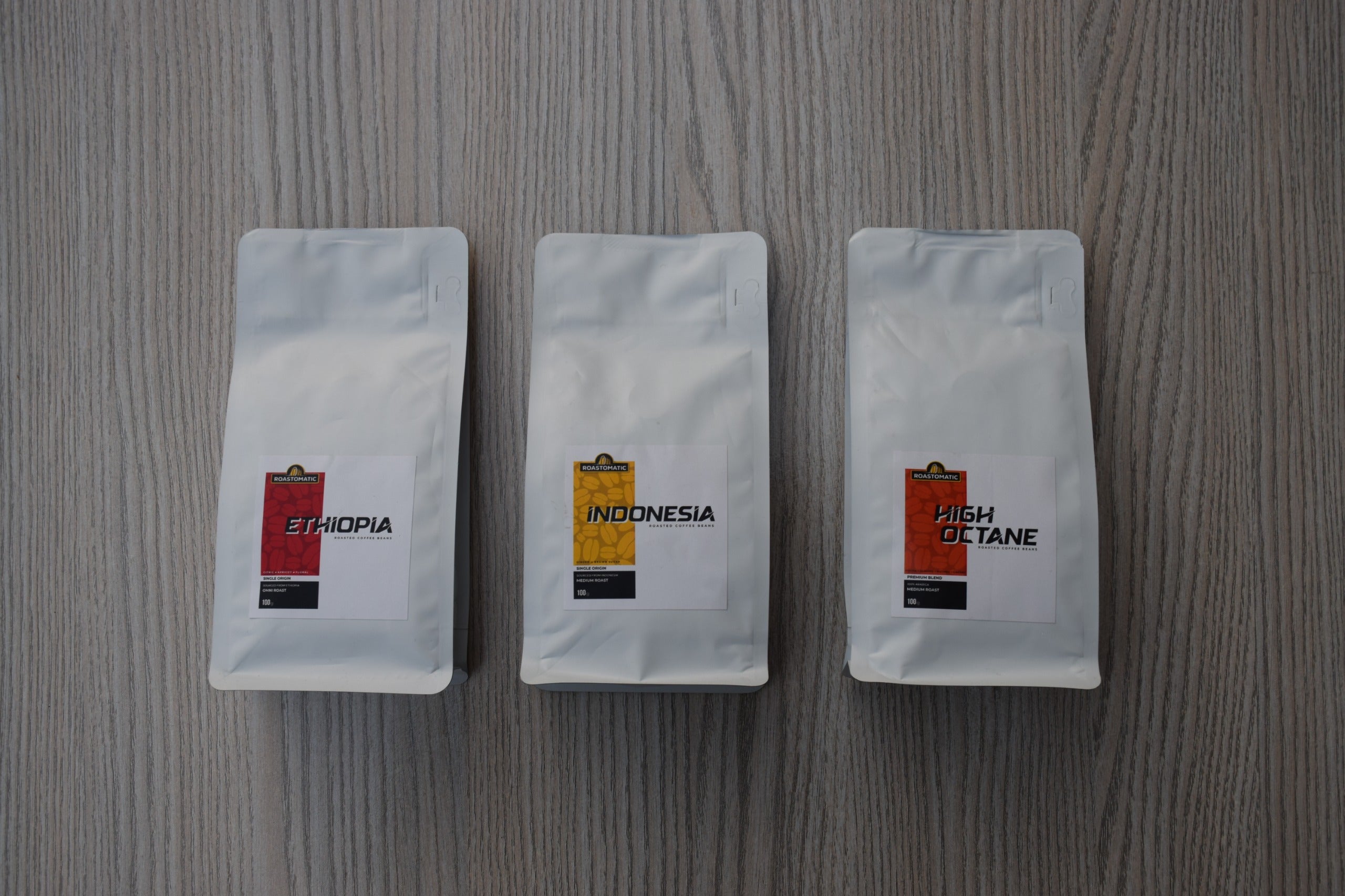 Roastomatic three white coffee bags labeled 'Ethiopia', 'Indonesia', and 'High Octane' on a wooden surface.