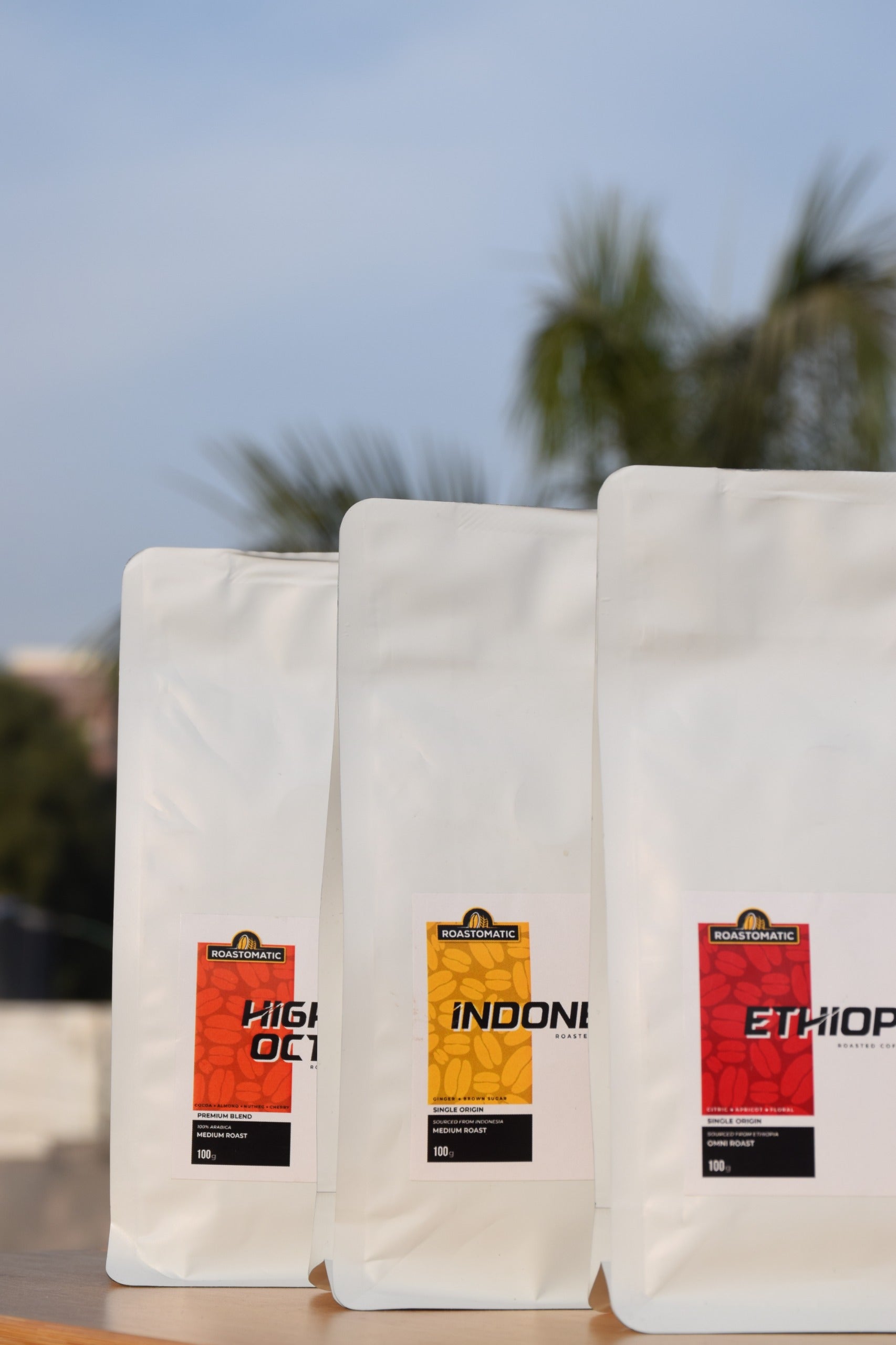 Roastomatic three coffee bags labeled 'High Altitude', 'Indoni', and 'Ethiopia' on a blurred natural background.